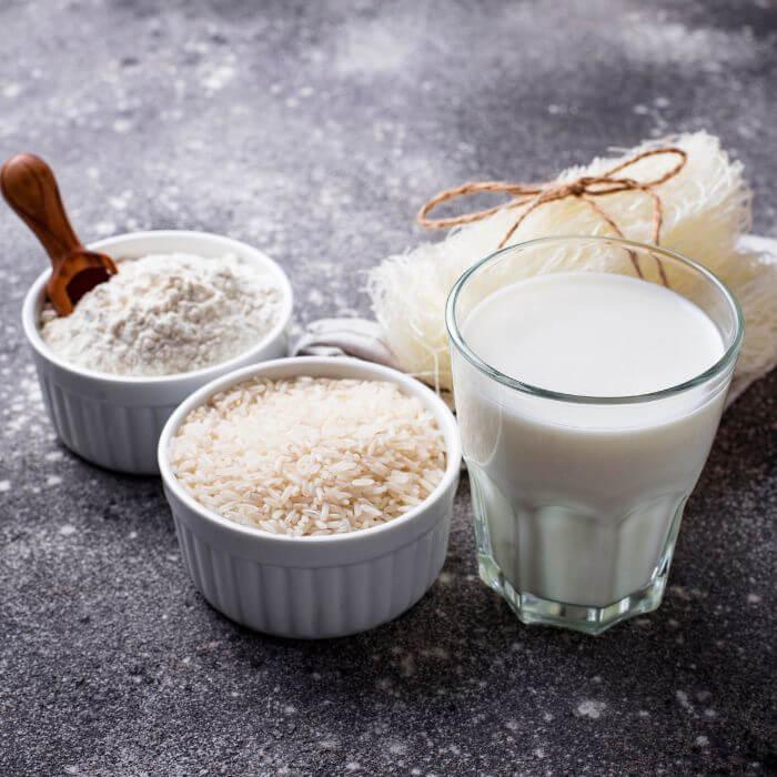 Glass of plant-based rice milk with raw rice, flour, and rice noodles on a gray surface