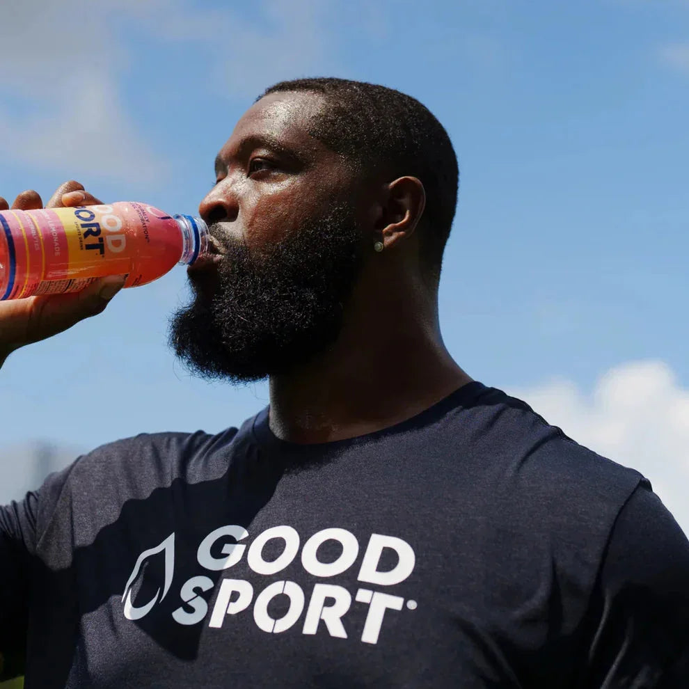 Man outdoors drinking GoodSport Strawberry Lemonade, wearing branded sports shirt, under blue sky