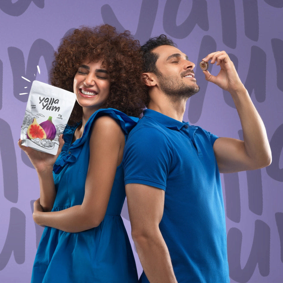 Smiling woman holding Yalla Yum fig snacks beside man eating a fig, plant-based food store UAE