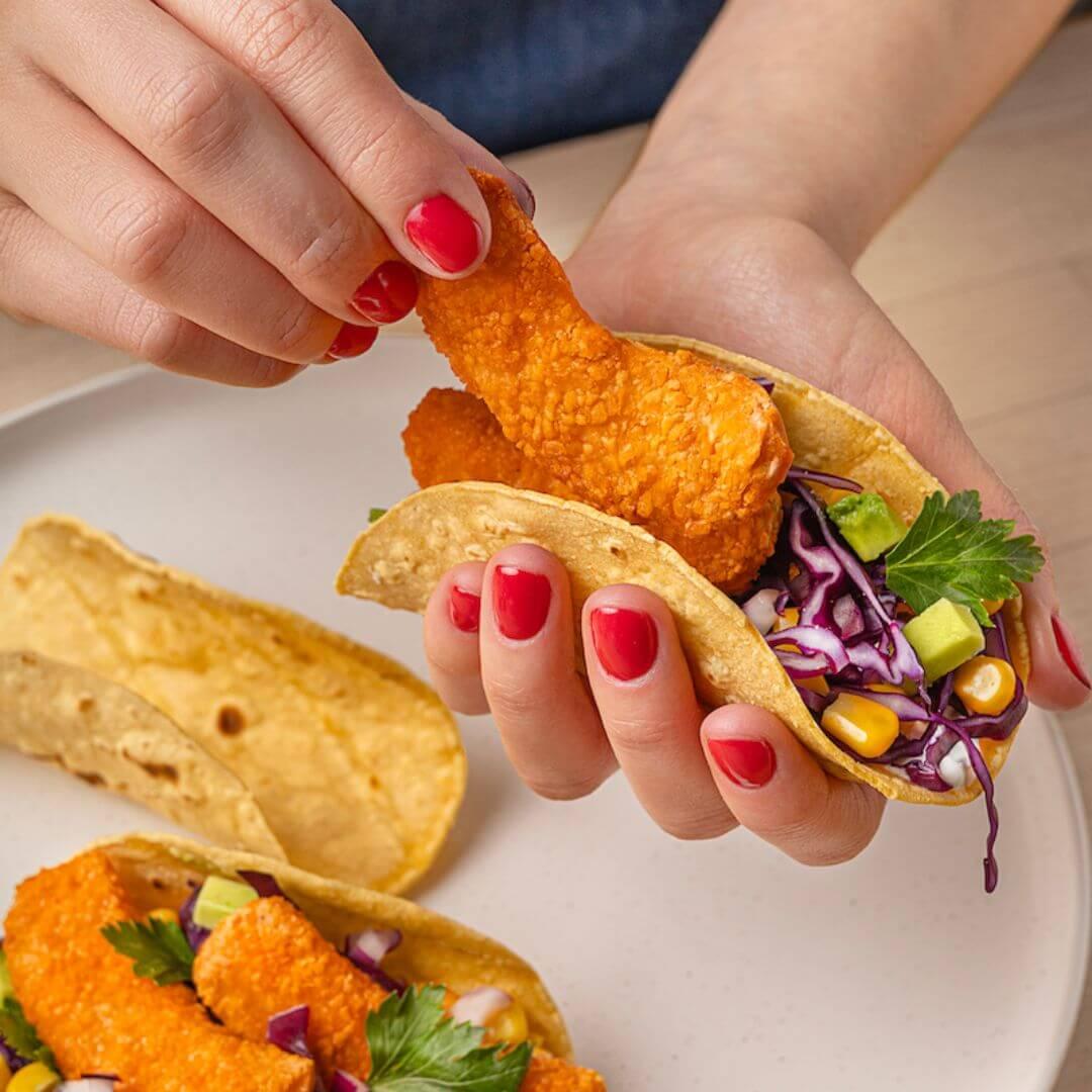 Hand assembling a plant-based taco with crispy vegan nuggets, corn, avocado, and cabbage on a plate
