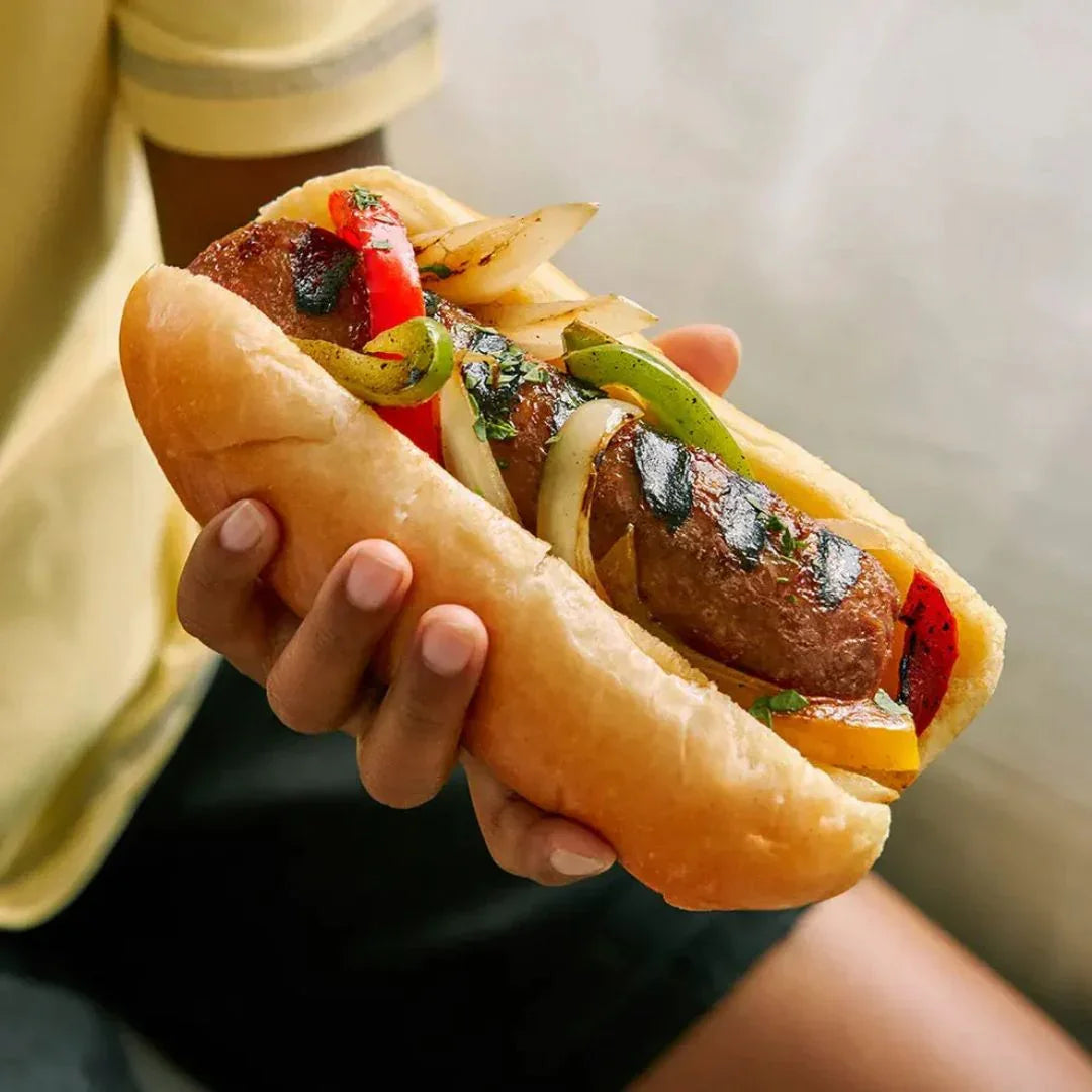 Hand holding a plant-based sausage sandwich with grilled peppers and onions in a bun
