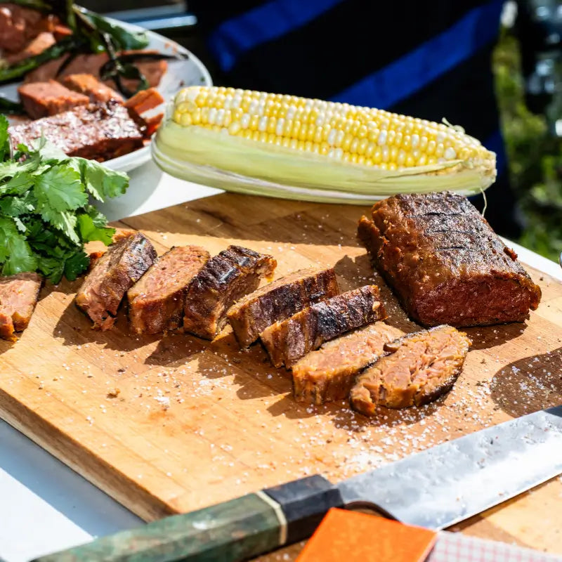 Sliced meaty meat on a wooden cutting board with an ear of corn and a knife, outdoors.