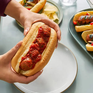 Hands holding a plant-based meatball sub with tomato sauce, surrounded by vegan hot dogs and chips.