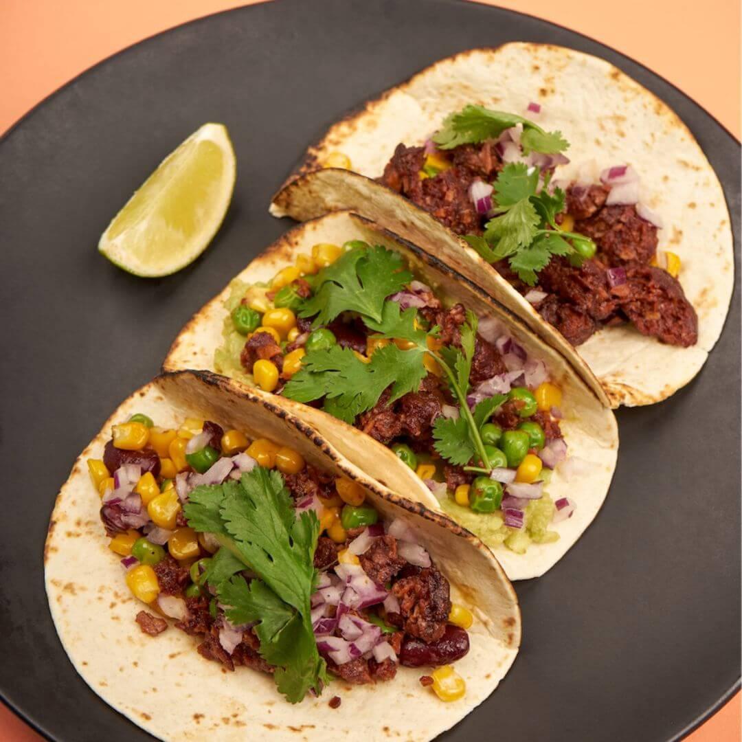 Three plant-based tacos with corn, beans, onions, and cilantro on a black plate, iPlantBased UAE