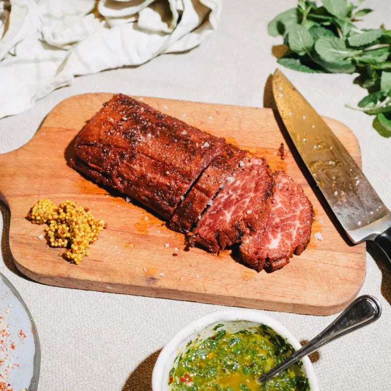 whole cut loin steak on a wooden cutting board with a knife, herbs, and a bowl of green sauce.